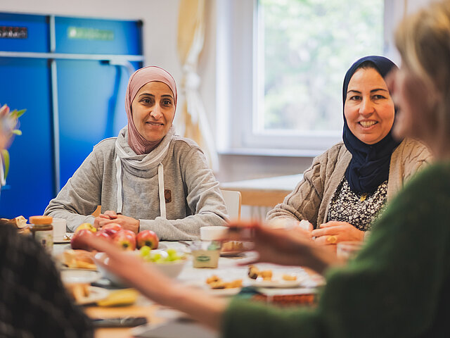 Vier Frauen sitzen um einen Tisch mit Essen, lächeln und unterhalten sich in einer hellen, ungezwungenen Umgebung.