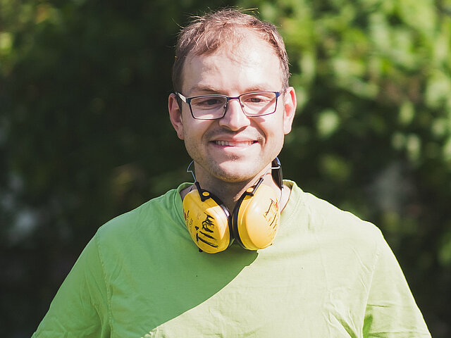 Mann mit Brille, grünem Hemd und Handschuhen, mit gelben Ohrenschützern um den Hals, steht im Freien.