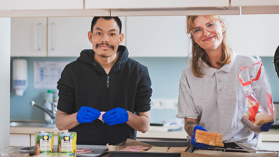Zwei Personen in einer Küche, die Essen zubereiten, lächelnd und mit blauen Handschuhen.