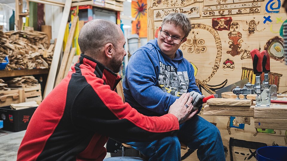 Ein Mann und ein lächelnder Junge arbeiten gemeinsam an einem Holzbearbeitungsprojekt in einer farbenfrohen, kreativen Werkstatt.