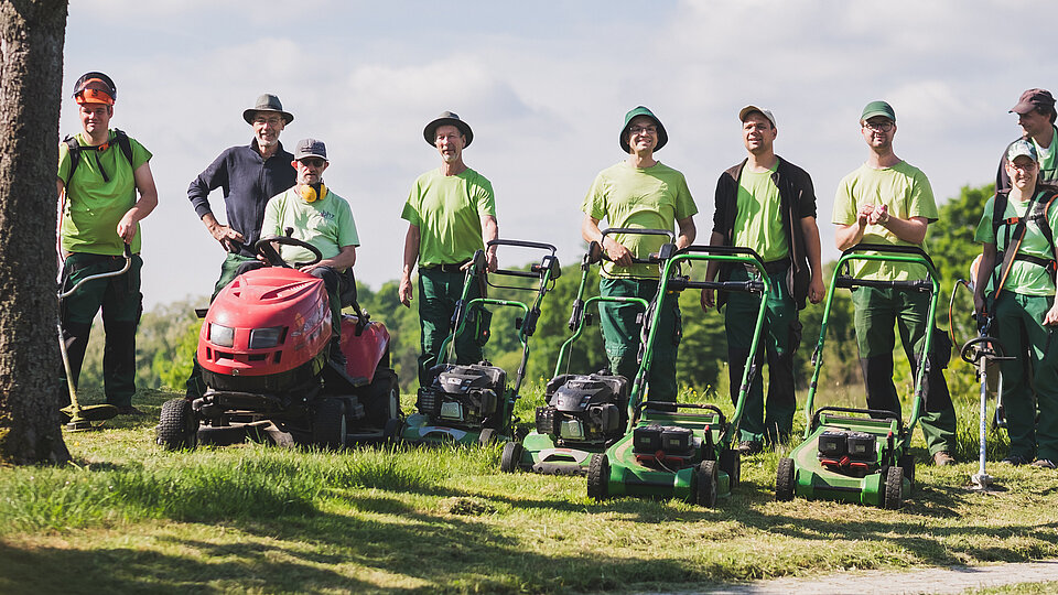 Eine Gruppe von Landschaftsgärtnern mit Rasenmähern und einem Aufsitzmäher steht an einem sonnigen Tag im Freien.